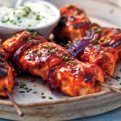 Buffalo chicken skewers topped with sauce and garnished with chives, served with blue cheese yogurt dip.