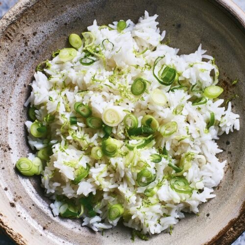 Bowl of coconut rice topped with chopped spring onions and lime wedges on the side.