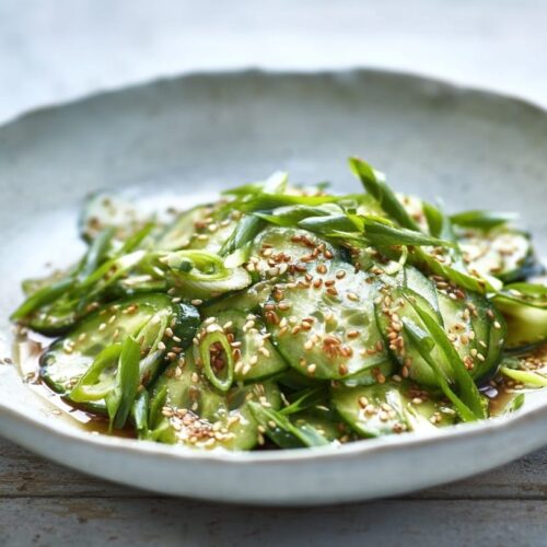 Sliced cucumbers and green onions in a shallow bowl with sesame seeds and dressing