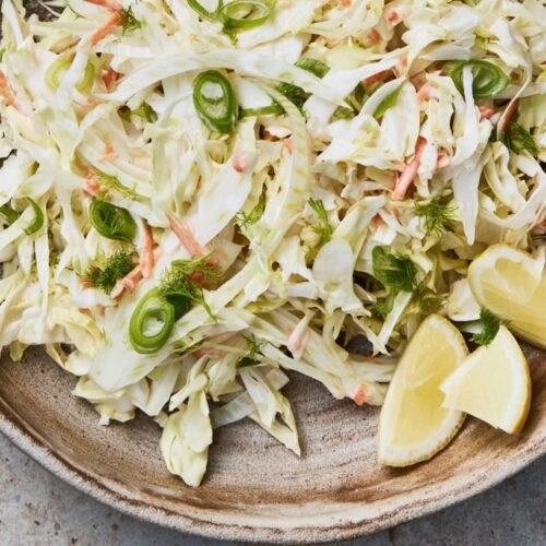 A bowl of fennel and cabbage slaw with shredded carrots and green onions, garnished with lemon wedges.