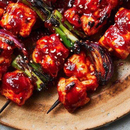 Grilled chicken skewers coated in red gochujang sauce with green onions and red onion on a wooden plate.