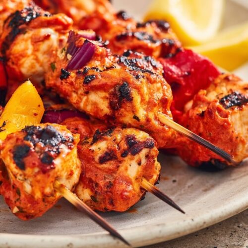 Grilled chicken skewers coated in harissa sauce, served with colorful bell peppers and lemon wedges.