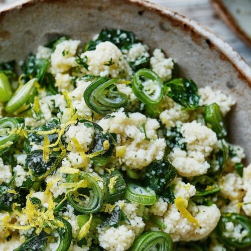 A bowl of herby couscous mixed with parsley, lemon zest, and green onions on a rustic surface.