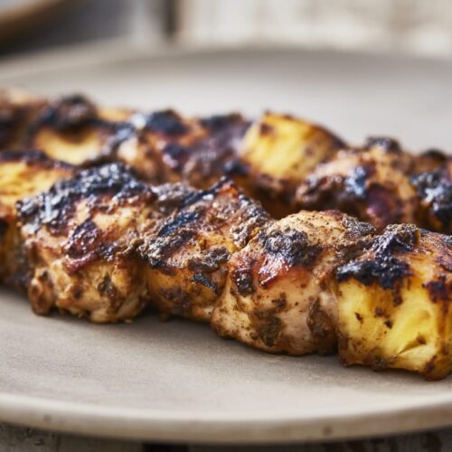 Grilled jerk chicken skewers with chunks of pineapple on a beige plate, showcasing a charred texture.