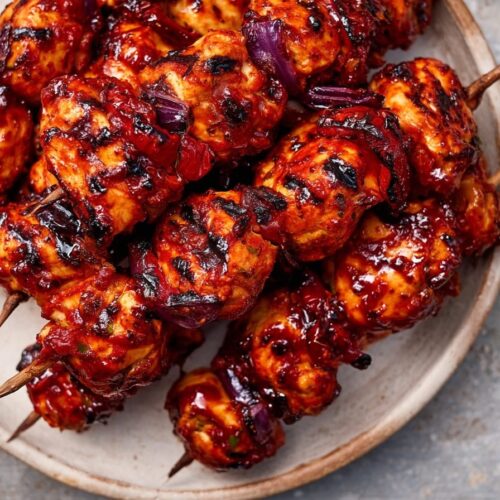 Grilled chicken skewers coated in spicy peri peri sauce with red peppers and lime wedges on a plate.