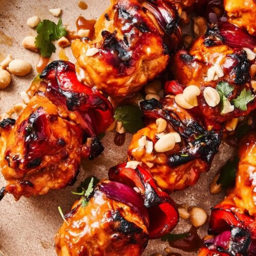 Grilled chicken skewers coated in Sriracha sauce, garnished with peanuts and herbs on a plate.