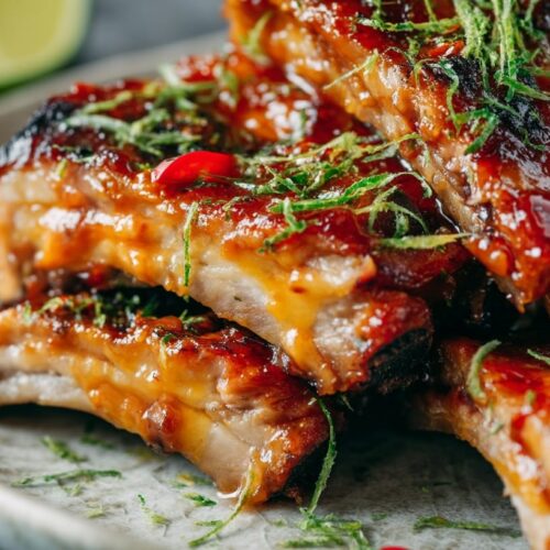 A plate of sticky mango habanero pork ribs garnished with herbs and red chili slices, with lime in the background.