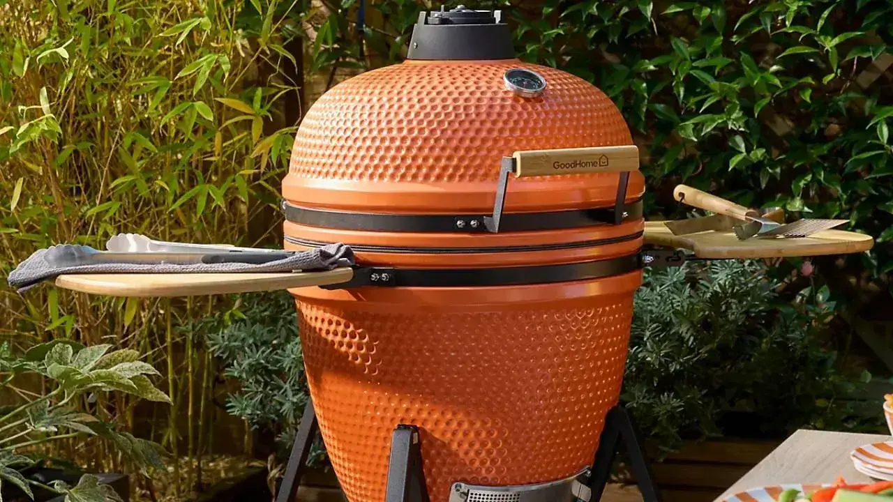 Orange ceramic BBQ grill with wooden side shelves and a black lid in a garden setting.