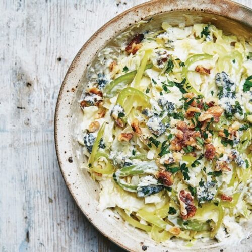 A bowl of coleslaw featuring blue cheese, celery, walnuts, and herbs on a wooden surface.