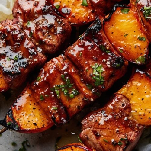 Grilled beef skewers with peaches, garnished with herbs and cracked black pepper on a plate.
