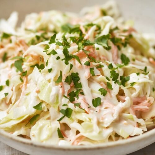 A bowl of creamy coleslaw made with shredded cabbage and carrots, garnished with chopped parsley.