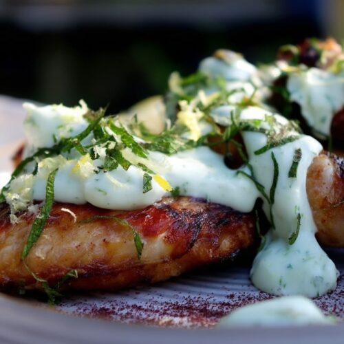 Grilled chicken wings topped with cucumber yoghurt sauce, mint, and lemon slices on a plate.