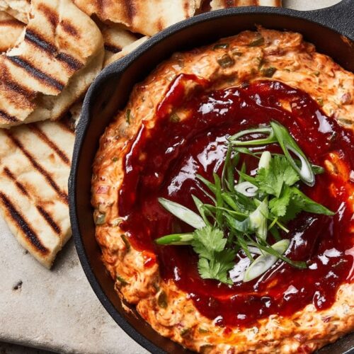 A creamy dip with a red sauce and green garnish in a cast iron skillet, served with grilled flatbread.