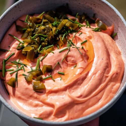 A bowl of pink dip topped with chopped jalape&ntilde;os and green herbs on a dark surface.