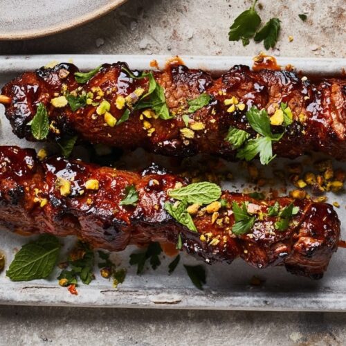 Two beef skewers topped with mint, pistachios, and a side of yogurt sauce.