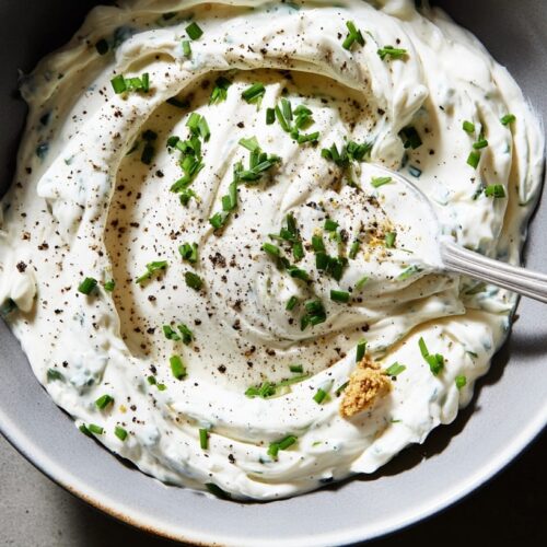 A creamy dip with chives and black pepper in a grey bowl on a stone surface.