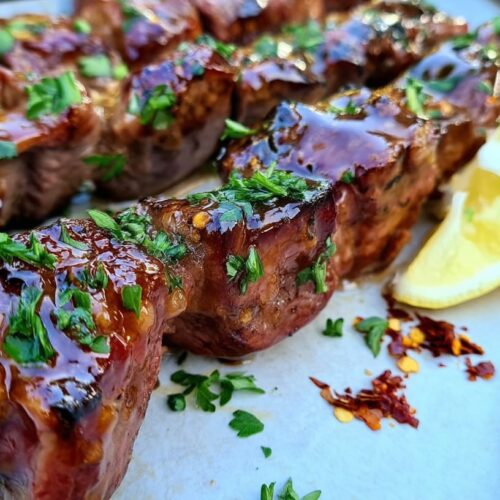 Grilled beef skewers glazed with honey garlic sauce, garnished with parsley and lemon slices.
