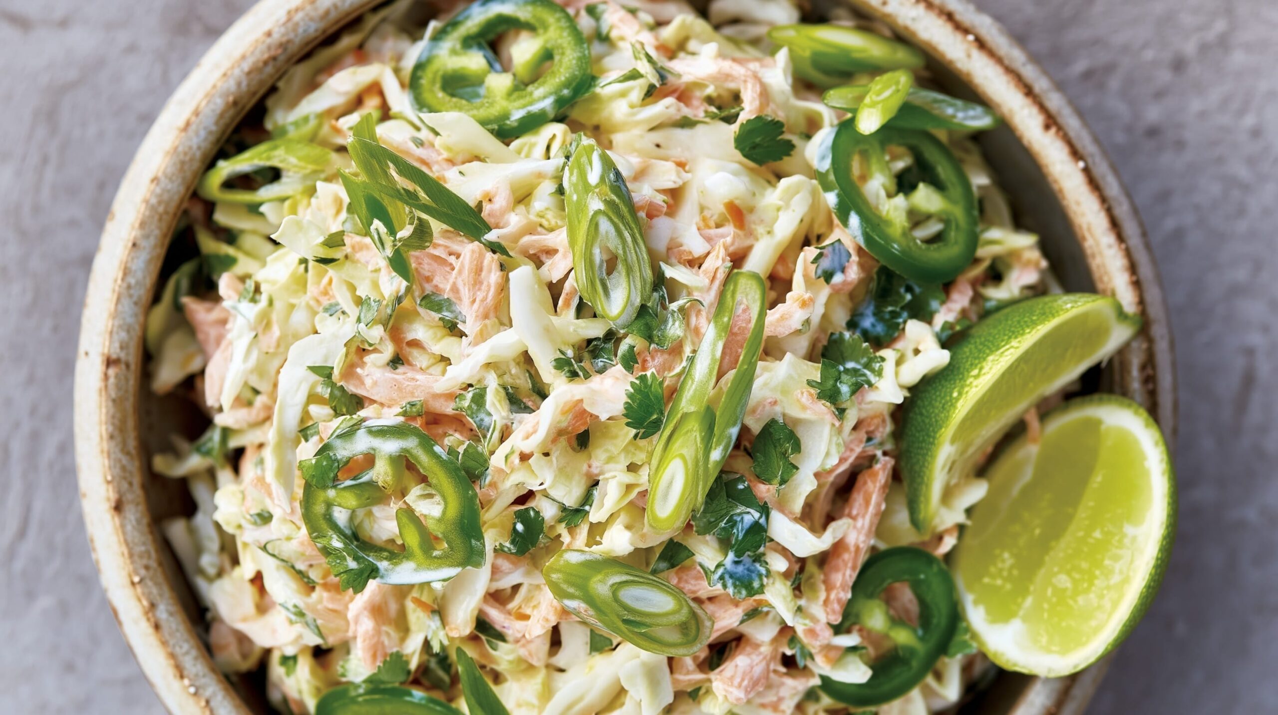A bowl of coleslaw with shredded cabbage, jalapeños, lime wedges, and coriander leaves.