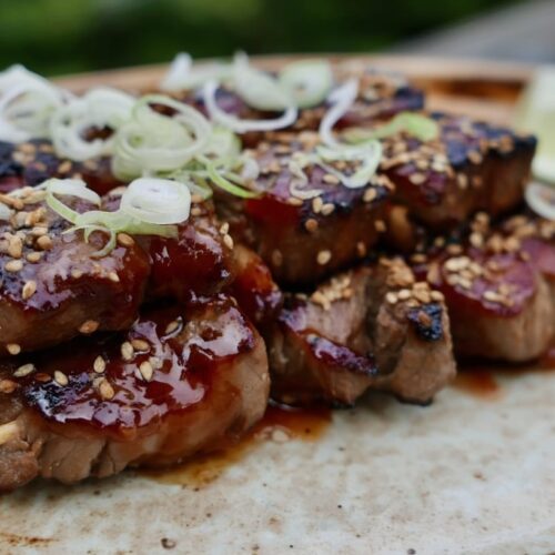 Three beef skewers topped with green onions and sesame seeds on a plate with lime slices.
