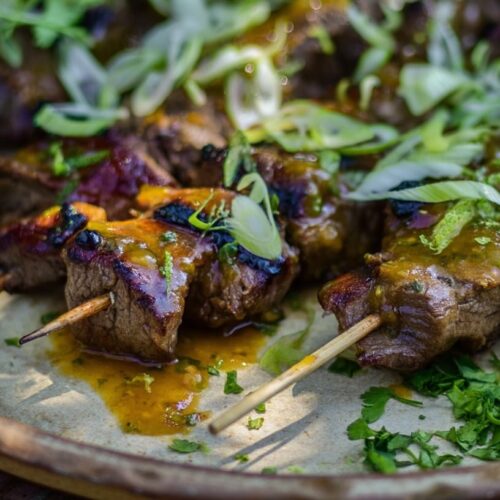 Grilled beef skewers topped with green onions, lime, and jalapeno slices on a plate.
