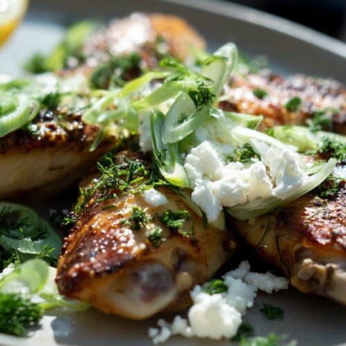 Golden brown chicken wings garnished with spring onion, feta cheese, and fresh herbs on a plate.