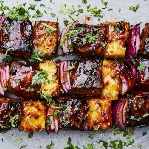 Grilled beef skewers with pineapple and red onion, garnished with chopped coriander on a plate.