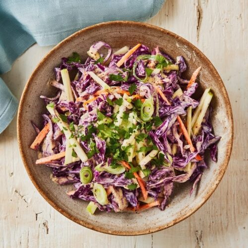 A bowl of red cabbage coleslaw with apple, mustard, and green onions on a wooden table.