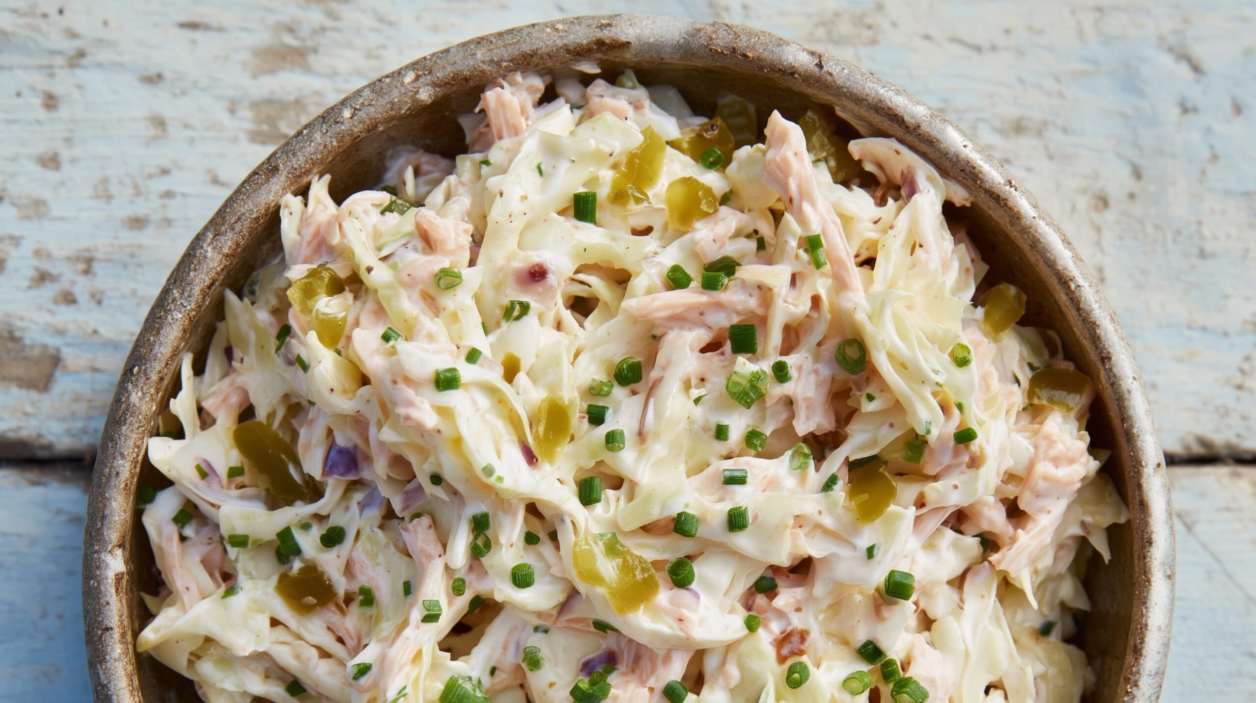 A bowl of coleslaw with shredded cabbage, smoked onion pickle, and chopped chives on a wooden surface.