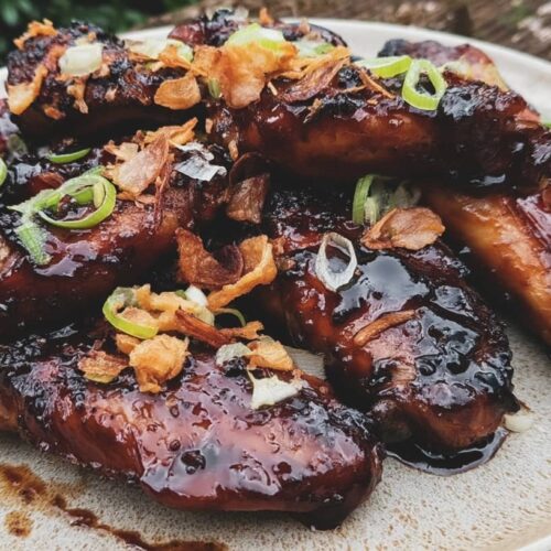A plate of chicken wings coated in a glossy black garlic soy treacle glaze, garnished with shallots.