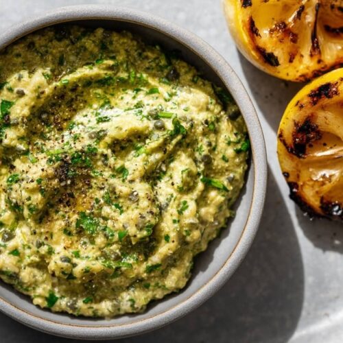 A grey bowl filled with a creamy dip alongside two charred lemon halves on a plate.