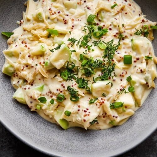 A creamy dip with green apple pieces, mustard seeds, and fresh herbs in a grey bowl.