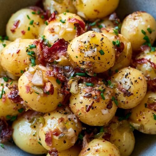 A bowl of mustard potato salad featuring small potatoes, bacon bits, chives, and pickles.