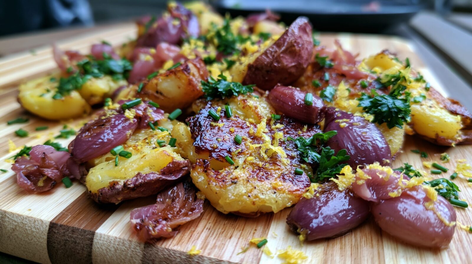 Smashed potatoes topped with pickled shallots, herbs, and lemon zest on a wooden board.