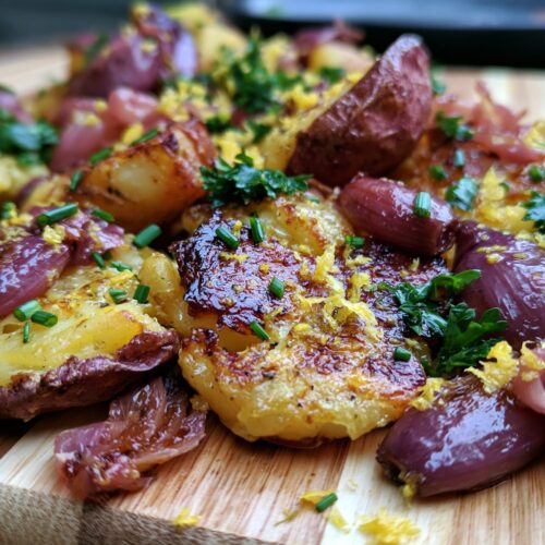 Smashed potatoes topped with pickled shallots, herbs, and lemon zest on a wooden board.